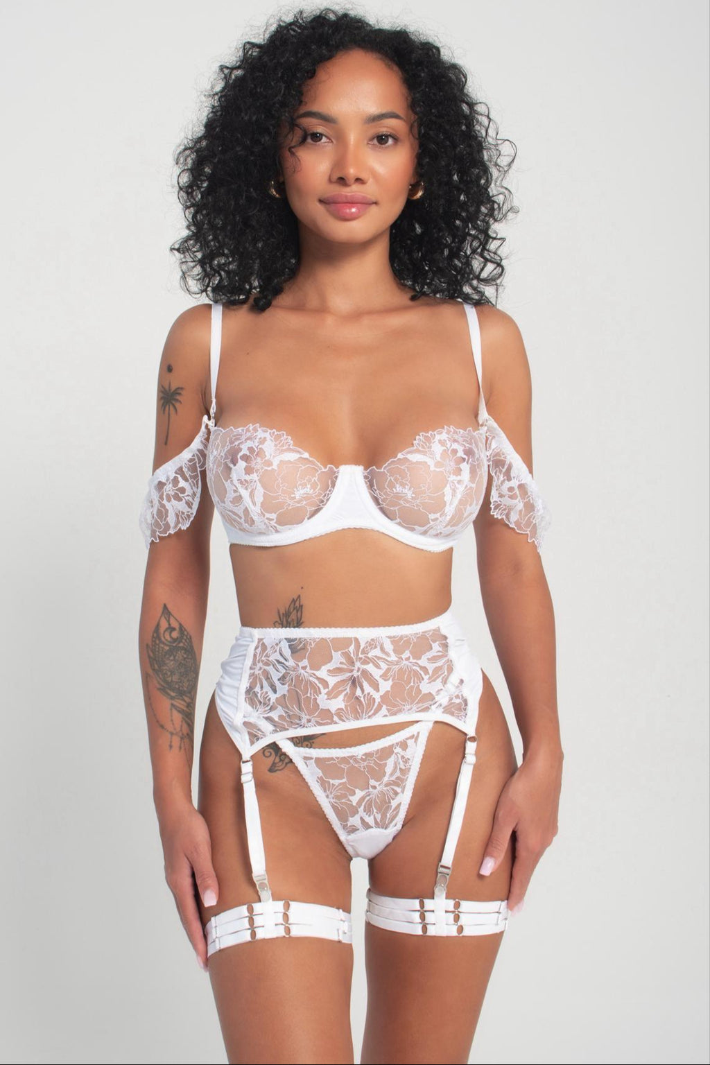 A model wearing a white lingerie set with intricate embroidery, including a bra, panties, garters, and a belt.