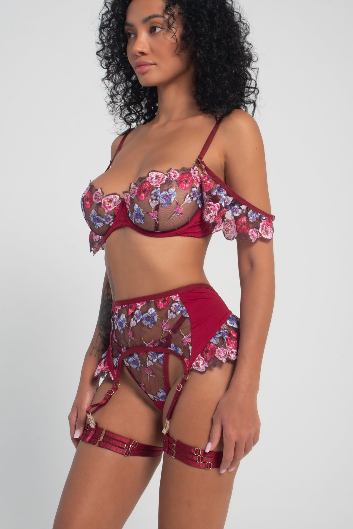 A model wearing a red and floral lace lingerie set with Brazilian and belt, including a bra, panties, and garter belt.