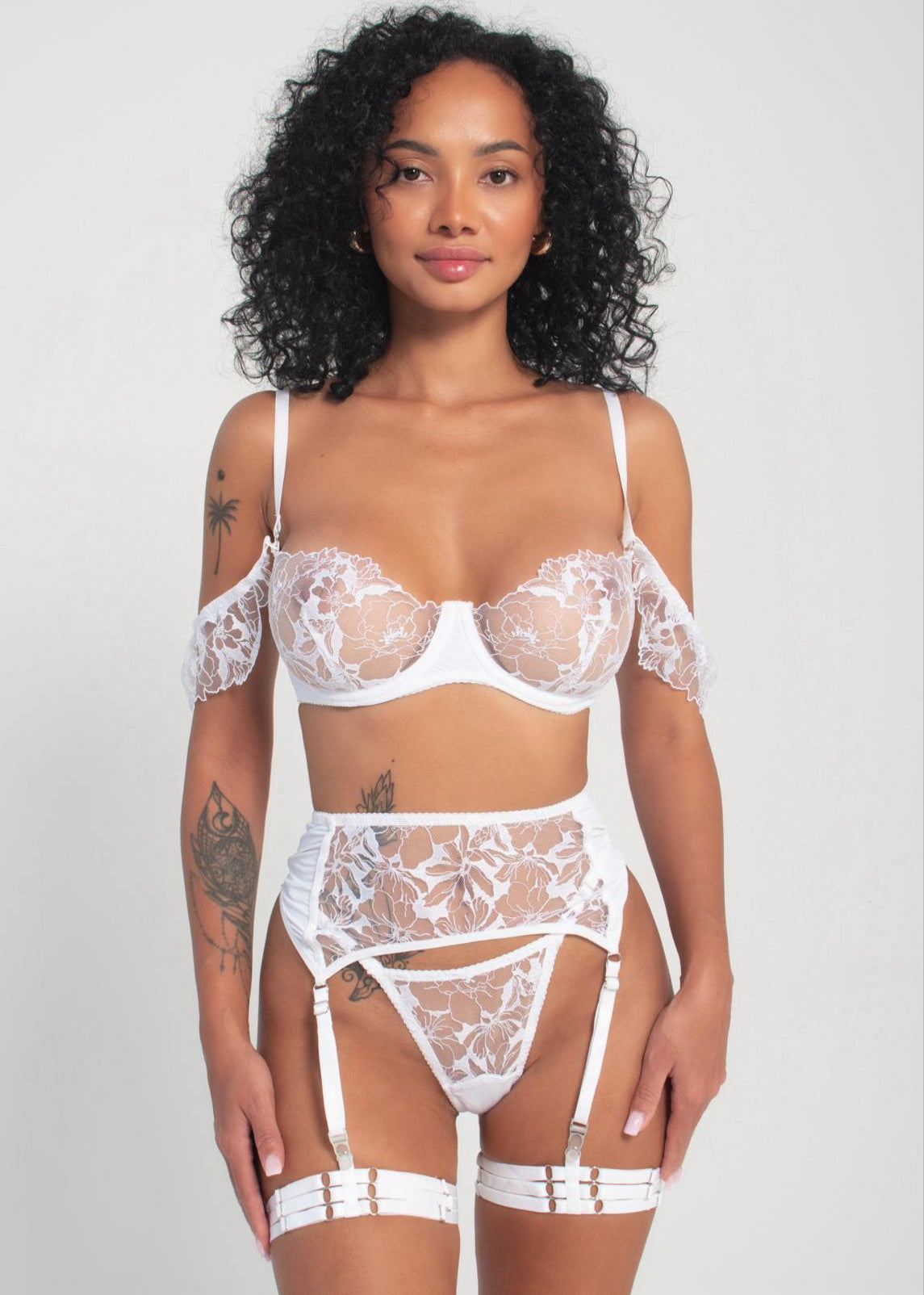 A model wearing a white lingerie set with intricate embroidery, including a bra, panties, garters, and a belt.